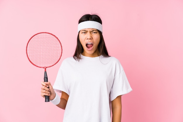 Young chinese woman playing badminton in a pink wall screaming very angry and aggressive.