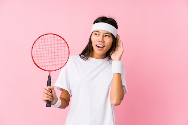 Young chinese woman playing badminton in a pink wall trying to listening a gossip.