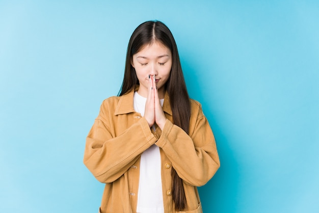 Young chinese woman posing in a blue wall isolated holding hands in pray near mouth, feels confident.