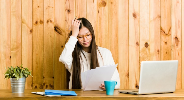 Photo young chinese woman studying on her desk forgetting something, slapping forehead with palm and closing eyes.