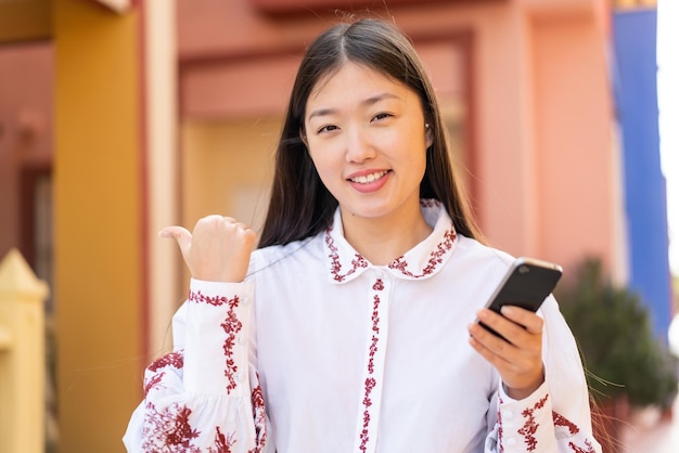 Young Chinese woman using mobile phone at outdoors pointing to the side to present a product