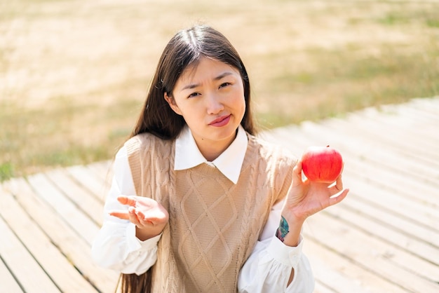 Young Chinese woman with an apple at outdoors making doubts gesture while lifting the shoulders