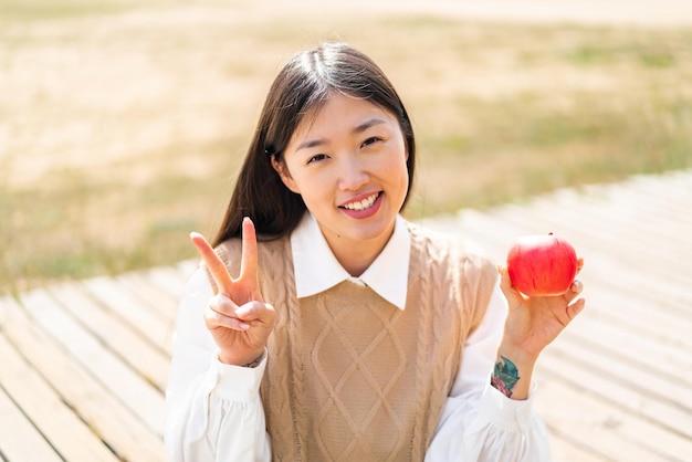 Young Chinese woman with an apple at outdoors smiling and showing victory sign