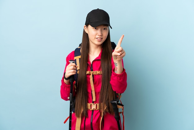 Photo young chinese woman with backpack and trekking poles over isolated blue wall showing and lifting a finger