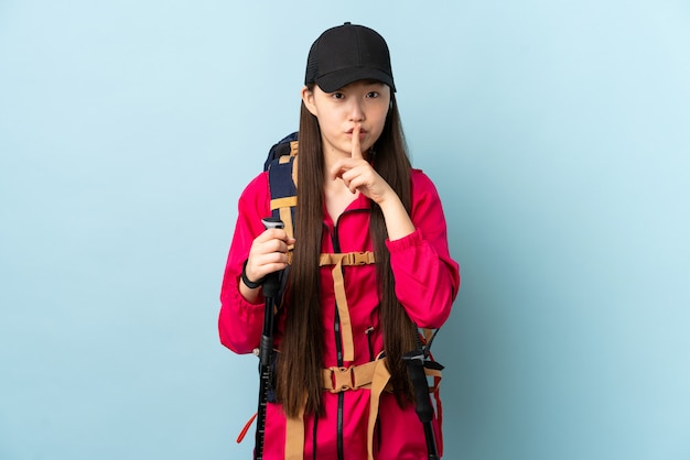 Young Chinese woman with backpack and trekking poles over isolated blue wall showing a sign of silence gesture putting finger in mouth