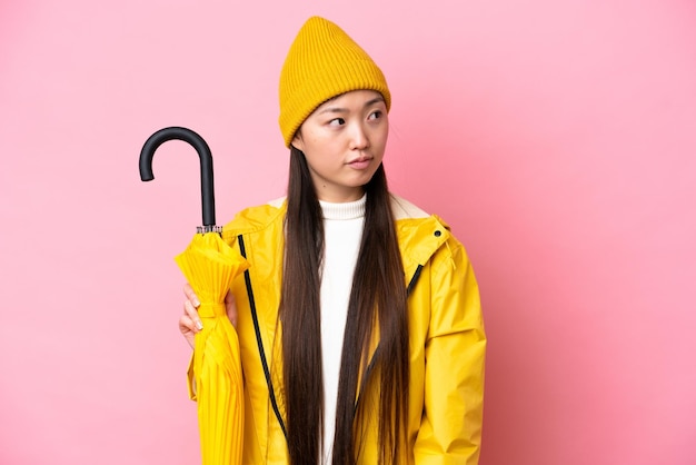 Young Chinese woman with rainproof coat and umbrella isolated on pink background looking to the side