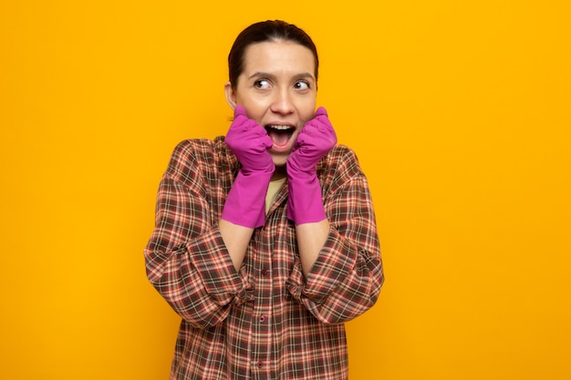 Young cleaning woman in plaid shirt in rubber gloves looking aside happy and excited holding hands together