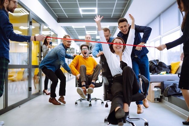 Young colleagues group having fun together riding on chairs in office