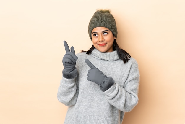 Young Colombian girl with winter hat on beige wall pointing with the index finger a great idea