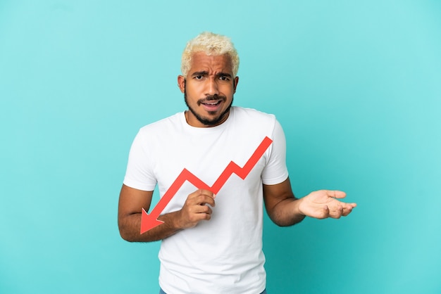 Young Colombian handsome man isolated on blue background holding a downward arrow and with sad expression