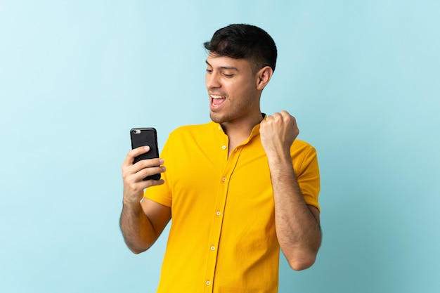 Young Colombian man using mobile phone isolated on blue celebrating a victory