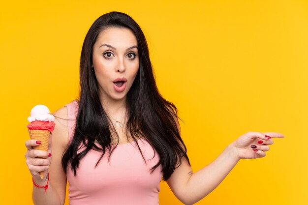 Young Colombian woman holding an cornet ice cream over isolated yellow wall surprised and pointing side