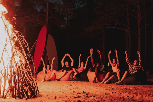 Young Company Sit In Front Of Bonfire On Beach.