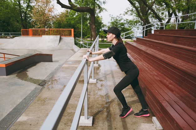 Young concentrated athletic beautiful brunette woman in black uniform, cap with earphones listening to music doing sport stretching exercises warming-up in city park outdoors