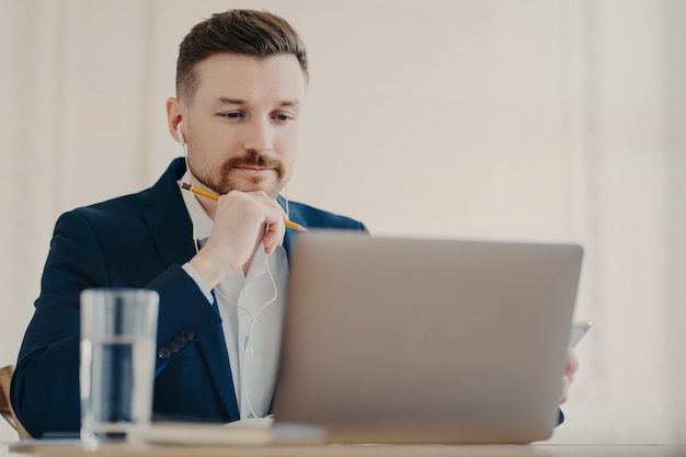 Young concentrated businessman or entrepreneur wearing earphones, holding pencil and looking at modern laptop screen while watching online business webinar. Working at desk in light office