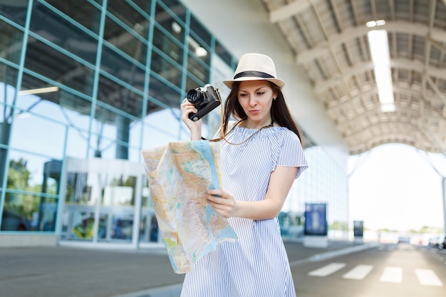 Young concerned traveler tourist woman in hat hold retro vintage photo camera, looking at paper map at international airport