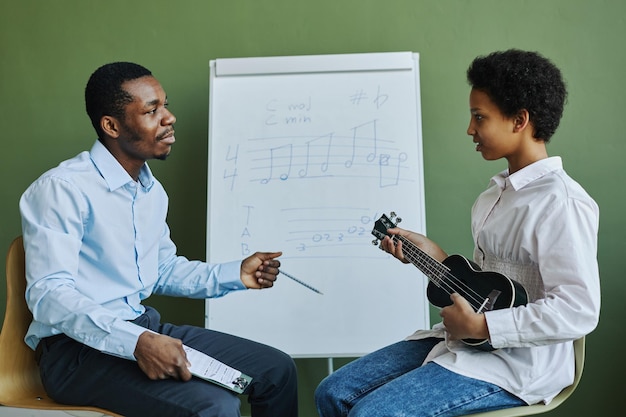 Young confident african american teacher answering questions of schoolgirl at lesson with ukulele wh