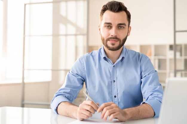 Young confident agent or consultant in blue shirt sitting by desk and looking at you ready to answer your questions