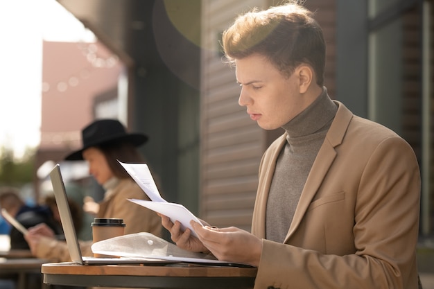 Young confident businessman in smart casualwear looking through business papers while sitting by table in front of laptop outdoors