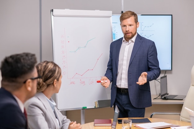 Young confident businessman with red highlighter discussing financial garph with colleagues