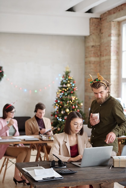 Young confident businessman with xmas headband pointing at laptop display while consulting female colleague about report for presentation