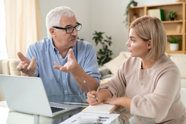 Young confident businesswoman looking at senior male colleague while discussing working points or plans at meeting in office