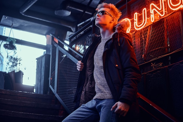 Young confident guy wearing coat with fur hood and sunglasses standing on the staircase to the underground nightclub, a backlit signboard in the background