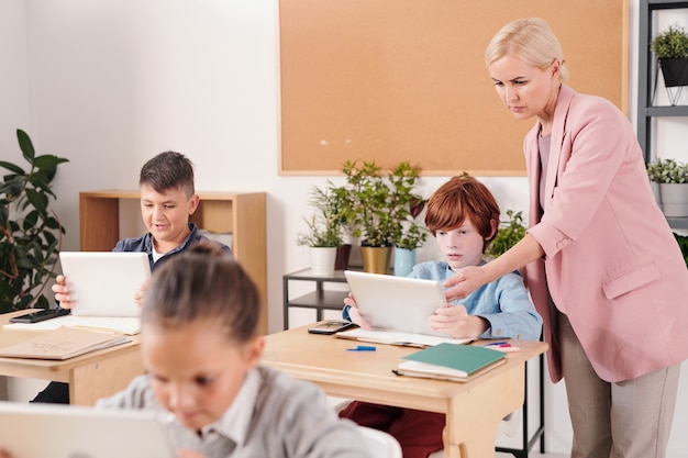 Young confident teacher in elegant suit consulting one of schoolkids with tablets at lesson during individual work with other pupils near by