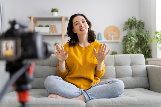 Young confident woman is talking on digital camera recording vlog sitting on sofa at home.