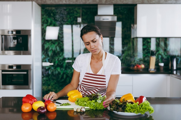 The young confused and thoughtful woman in apron decides what to cook in the kitchen