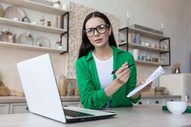 A young confused woman sits at home in the kitchen with a laptop jots down the family budget in a