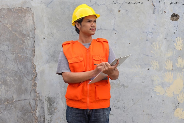 Young construction worker in yellow helmet and orange waistcoat holding a clipboard and looking away