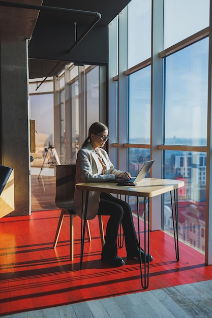 Young content worker in casual clothes sitting at a desk and using a netbook while working on the background of a spacious office Business woman in the office works sitting near the window