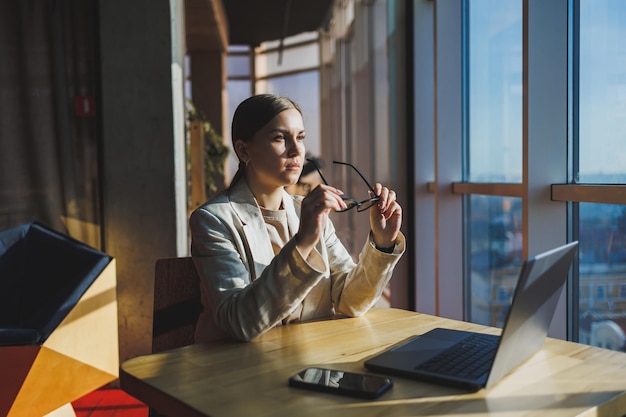 Photo young content worker in casual clothes sitting at a desk and using a netbook while working on the background of a spacious office business woman in the office works sitting near the window