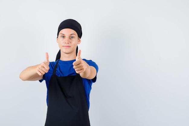 Young  cook showing thumbs up in t-shirt, apron and looking glad. front view.