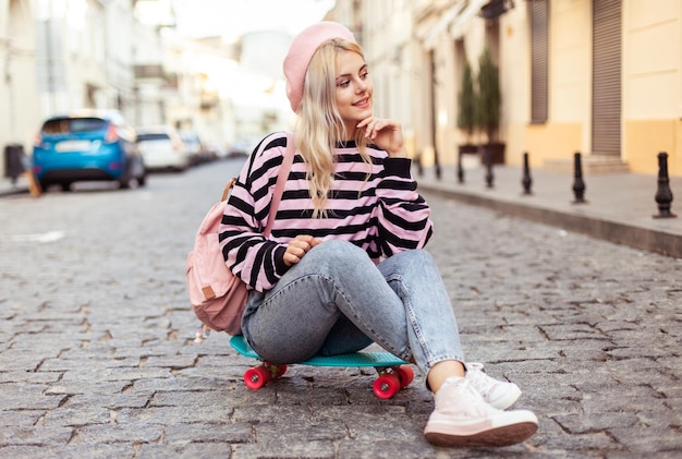 Young cool girl in a beret with a backpack sits on skateboard on European street