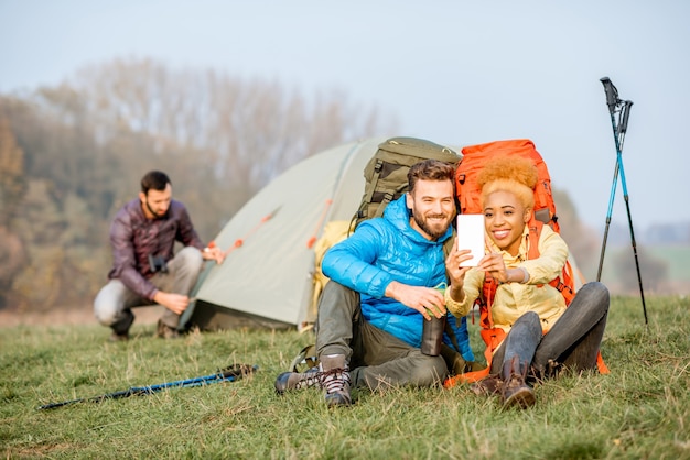 Young couple in colorful jackets with backpacks having fun with smart phone sitting on the green lawn near the camping place