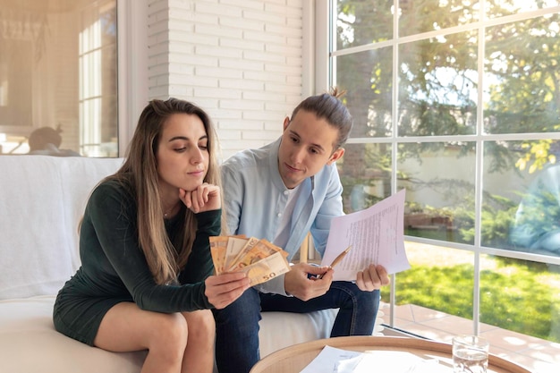 Young couple concern about money left for the month looking euro bills in hands sitting in the couch