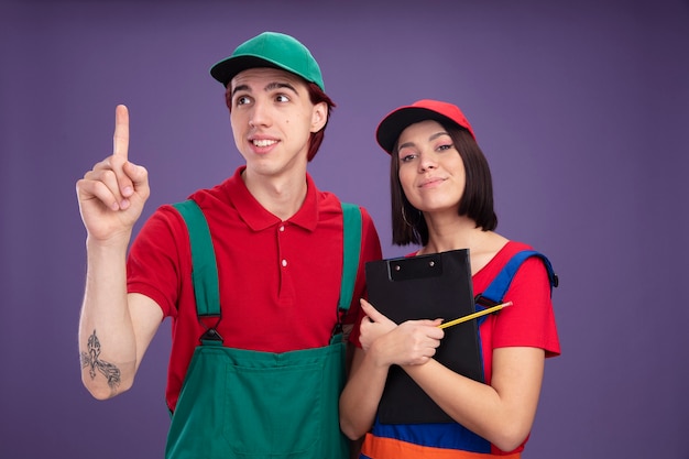 Young couple in construction worker uniform and cap girl holding pencil and clipboard smiling guy looking at side pointing up pleased girl hugging clipboard  isolated