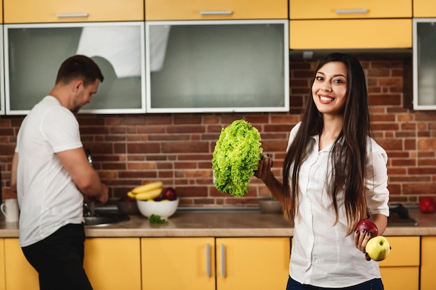 Young couple cooking in the kitchen. Woman smiling, holding lettuce and apples. The man in the background. Healthy Eating