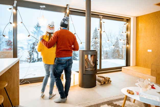 Young couple dressed in bright sweaters enjoying landscape view on the mountains standing together in the modern house during winter time