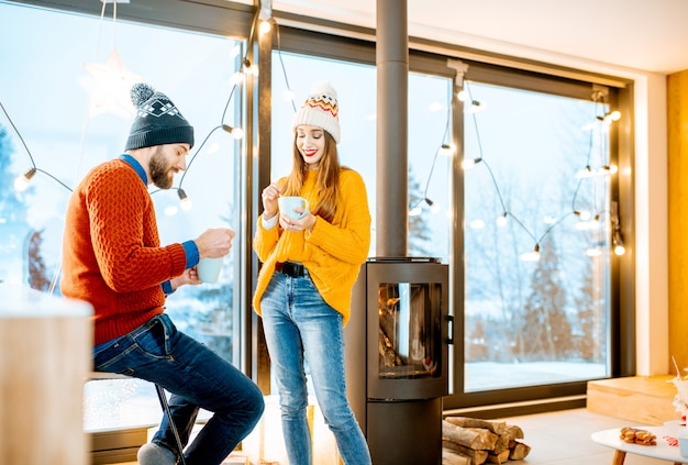 Young couple dressed in bright sweaters and hats standing together with hot drinks near the fireplace in the modern house durnig winter time