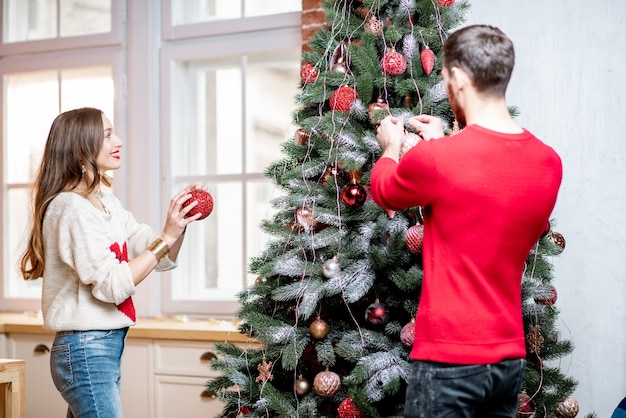 Young couple dressed in sweaters decorating Christmas tree preparing for a New Year holidays in their home