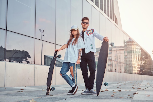 Young couple of dressed in trendy clothes posing with skateboards near skyscraper.