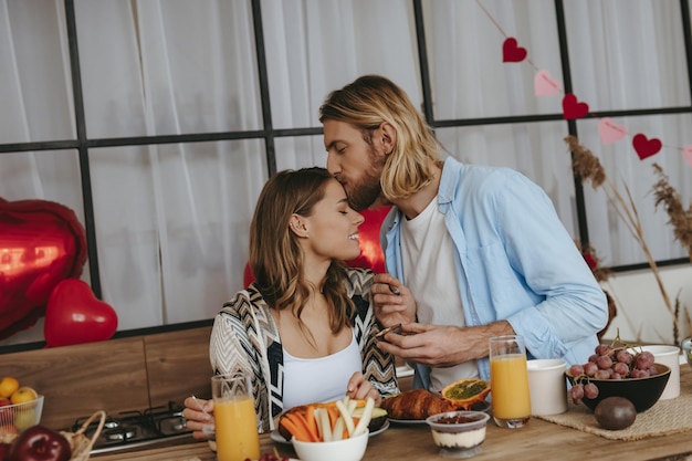 Young couple enjoying breakfast at the kitchen together with red heart shape balloons on background