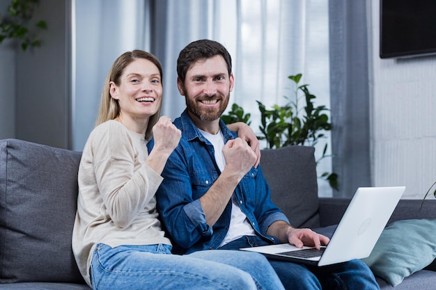 Young couple family husband and wife sitting on sofa at home using laptop winning something getting good news rejoicing smiling hugging