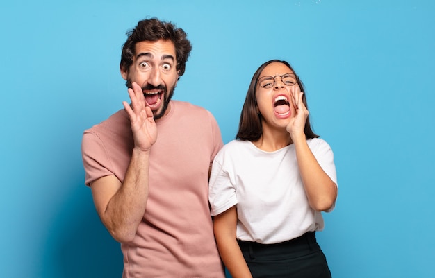 Young couple feeling happy, excited and positive, giving a big shout out with hands next to mouth, calling out