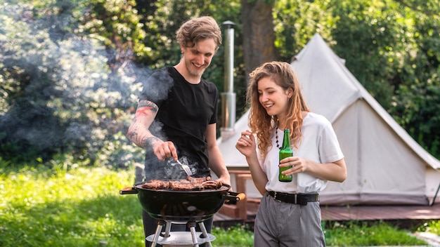 Young couple frying meat on the grill and drinking beer. Greenery around. Glamping