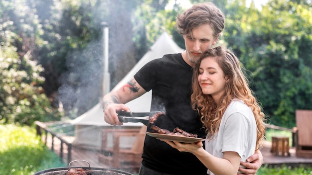 Young couple frying meat on the grill and putting it on the plate. Greenery around. Glamping