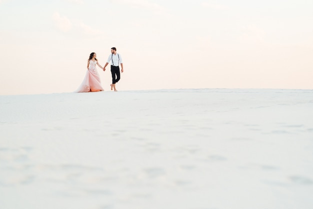 Young couple a guy in black breeches and a girl in a pink dress are walking along the white sand of the desert